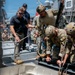 Military and civilian personnel are gathered on the deck of a naval vessel, working with ropes and rigging equipment. Several individuals wearing tactical helmets and camouflage uniforms appear to be conducting a training exercise or operational preparation involving rope systems. A civilian in a black polo shirt and sunglasses is actively participating alongside uniformed military members.