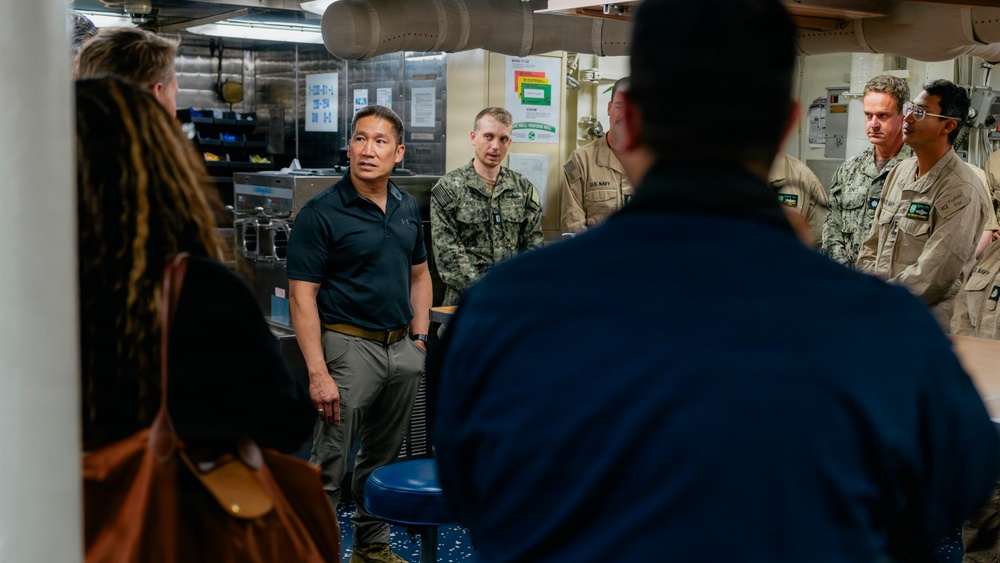 A group of individuals, including several U.S. Navy personnel in camouflage uniforms and civilians in casual attire, are gathered in what appears to be a shipboard galley or mess facility. A man in a black polo shirt and khaki pants appears to be the focal point of a briefing or discussion. The setting suggests an official visit or tour of a naval vessel.