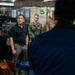 A group of individuals, including several U.S. Navy personnel in camouflage uniforms and civilians in casual attire, are gathered in what appears to be a shipboard galley or mess facility. A man in a black polo shirt and khaki pants appears to be the focal point of a briefing or discussion. The setting suggests an official visit or tour of a naval vessel.