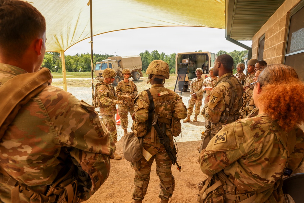 A group of U.S. Army soldiers in OCP (Operational Camouflage Pattern) uniforms are gathered under a tan shade canopy at what appears to be a field training exercise or military installation. A briefing or instruction session appears to be taking place, with one soldier gesturing while addressing the group. Military tactical vehicles are visible in the background.