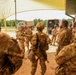 A group of U.S. Army soldiers in OCP (Operational Camouflage Pattern) uniforms are gathered under a tan shade canopy at what appears to be a field training exercise or military installation. A briefing or instruction session appears to be taking place, with one soldier gesturing while addressing the group. Military tactical vehicles are visible in the background.