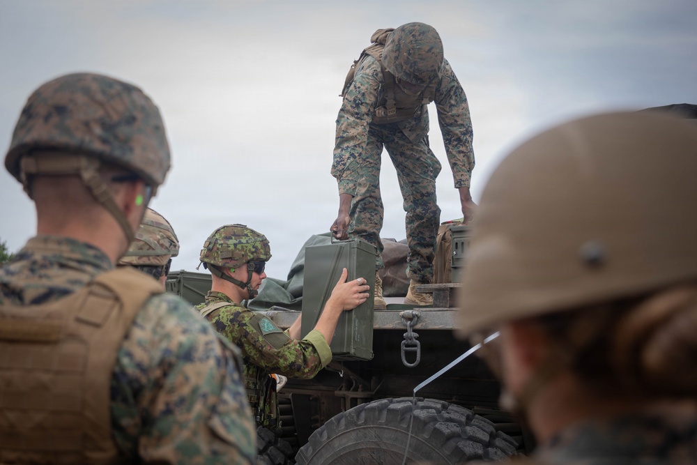 Military personnel in mixed camouflage uniforms are unloading or transferring equipment from a military vehicle. At least two distinct camouflage patterns are visible, suggesting a multinational or joint training exercise. Soldiers are passing what appears to be an ammunition can or military storage case from the truck bed to ground level.