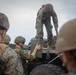 Military personnel in mixed camouflage uniforms are unloading or transferring equipment from a military vehicle. At least two distinct camouflage patterns are visible, suggesting a multinational or joint training exercise. Soldiers are passing what appears to be an ammunition can or military storage case from the truck bed to ground level.