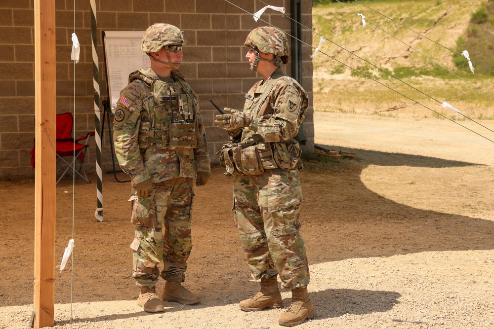 Two U.S. Army soldiers in Operational Camouflage Pattern (OCP) uniforms are engaged in conversation at what appears to be a military training range or field exercise area. Both soldiers are wearing combat helmets and tactical vests with equipment. The setting includes a cinder block building, a wooden post with range markers, and wire lines with white flags in the background.