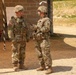 Two U.S. Army soldiers in Operational Camouflage Pattern (OCP) uniforms are engaged in conversation at what appears to be a military training range or field exercise area. Both soldiers are wearing combat helmets and tactical vests with equipment. The setting includes a cinder block building, a wooden post with range markers, and wire lines with white flags in the background.