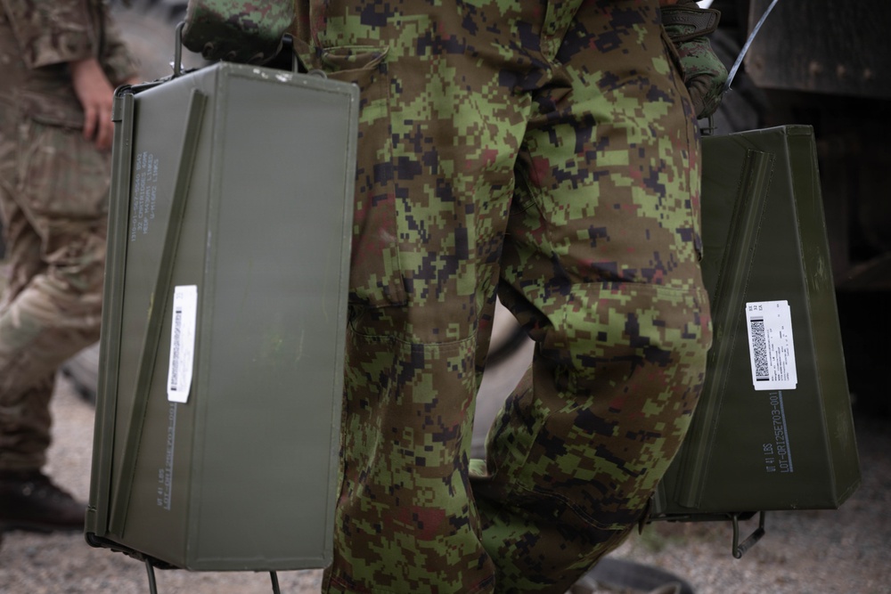 A soldier in digital camouflage uniform is carrying two olive-drab military ammunition cans, one in each hand. The cans bear barcode labels and stenciled markings typical of military ordnance containers. A second soldier in a different camouflage pattern is partially visible in the background to the left.