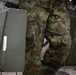 A soldier in digital camouflage uniform is carrying two olive-drab military ammunition cans, one in each hand. The cans bear barcode labels and stenciled markings typical of military ordnance containers. A second soldier in a different camouflage pattern is partially visible in the background to the left.