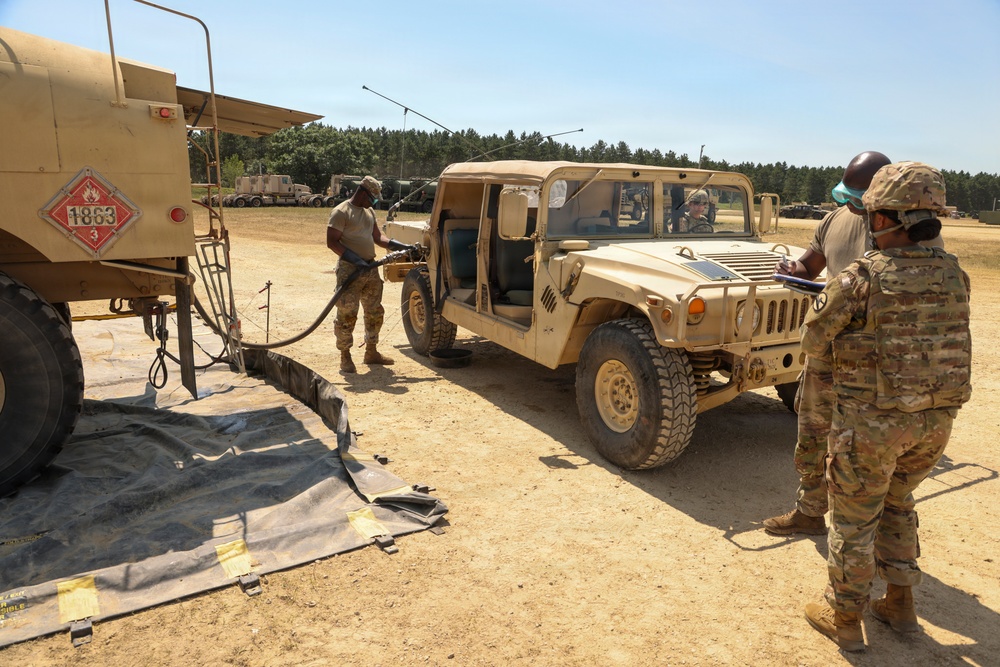 Military personnel in OCP (Operational Camouflage Pattern) uniforms are conducting a field refueling operation at what appears to be a military training area. A tan HMMWV (Humvee) is being refueled from a fuel tanker truck marked with a hazmat placard (UN 1863, flammable liquid). One soldier holds the fuel nozzle while another records information on a clipboard, and a third occupant remains inside the vehicle.