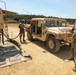 Military personnel in OCP (Operational Camouflage Pattern) uniforms are conducting a field refueling operation at what appears to be a military training area. A tan HMMWV (Humvee) is being refueled from a fuel tanker truck marked with a hazmat placard (UN 1863, flammable liquid). One soldier holds the fuel nozzle while another records information on a clipboard, and a third occupant remains inside the vehicle.