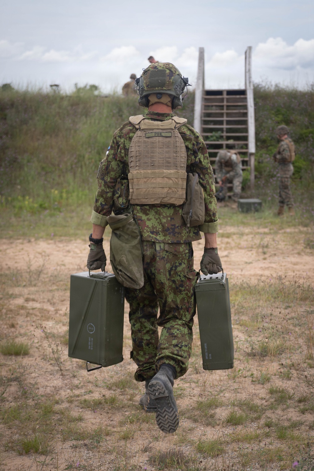 A soldier wearing Estonian military camouflage (EstCam pattern) and a tan plate carrier vest with the name tape 'PIHL' is walking away from the camera carrying two green military ammunition cans and a green bag. The setting appears to be a military training range or exercise area with sandy terrain, vegetation, and a wooden staircase structure in the background. Several other soldiers in mixed uniforms are visible in the background.