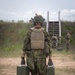 A soldier wearing Estonian military camouflage (EstCam pattern) and a tan plate carrier vest with the name tape 'PIHL' is walking away from the camera carrying two green military ammunition cans and a green bag. The setting appears to be a military training range or exercise area with sandy terrain, vegetation, and a wooden staircase structure in the background. Several other soldiers in mixed uniforms are visible in the background.