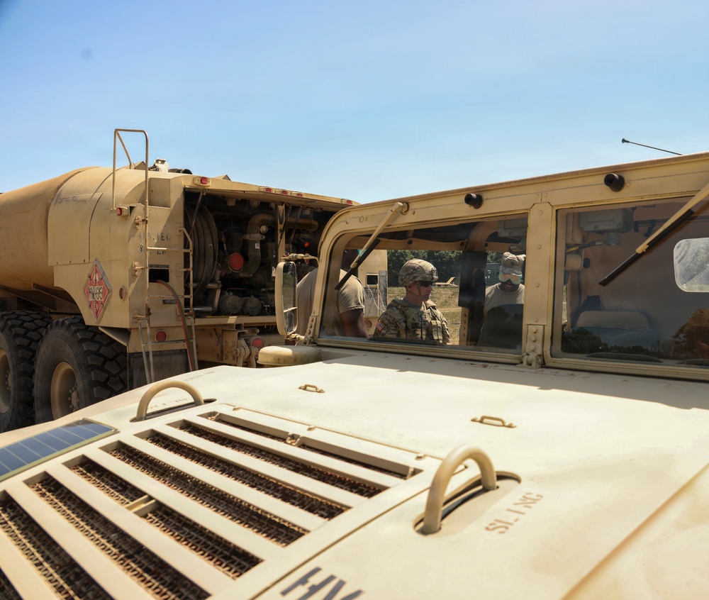 The image shows military personnel operating near two desert-tan painted military vehicles — a HMMWV (Humvee) in the foreground and a large fuel/tanker truck to the left. At least two soldiers in combat uniforms and helmets are visible through the Humvee's windshield area. The scene appears to be a field refueling or logistics operation conducted in an open outdoor area under clear skies.