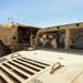 The image shows military personnel operating near two desert-tan painted military vehicles — a HMMWV (Humvee) in the foreground and a large fuel/tanker truck to the left. At least two soldiers in combat uniforms and helmets are visible through the Humvee's windshield area. The scene appears to be a field refueling or logistics operation conducted in an open outdoor area under clear skies.