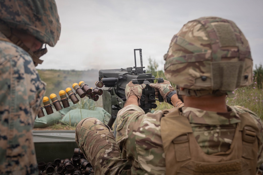 Two military personnel are operating what appears to be an Mk 19 automatic grenade launcher during a training exercise or live-fire event. One soldier wearing MultiCam/OCP pattern uniform is operating the weapon, while another in MARPAT (Marine digital) uniform observes from the left. A belt of 40mm grenades with yellow-tipped rounds is visible, and smoke can be seen downrange.