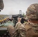 Two military personnel are operating what appears to be an Mk 19 automatic grenade launcher during a training exercise or live-fire event. One soldier wearing MultiCam/OCP pattern uniform is operating the weapon, while another in MARPAT (Marine digital) uniform observes from the left. A belt of 40mm grenades with yellow-tipped rounds is visible, and smoke can be seen downrange.
