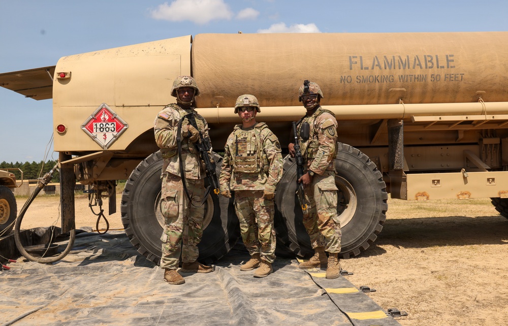 Three U.S. Army soldiers in MultiCam (OCP) camouflage uniforms and combat gear pose in front of a tan military fuel tanker truck. The tanker displays a hazmat placard with UN number 1863 (aviation fuel) and bears the warning 'FLAMMABLE - NO SMOKING WITHIN 50 FEET'. The setting appears to be an outdoor military training or operational area with sandy terrain.