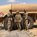 Three U.S. Army soldiers in MultiCam (OCP) camouflage uniforms and combat gear pose in front of a tan military fuel tanker truck. The tanker displays a hazmat placard with UN number 1863 (aviation fuel) and bears the warning 'FLAMMABLE - NO SMOKING WITHIN 50 FEET'. The setting appears to be an outdoor military training or operational area with sandy terrain.