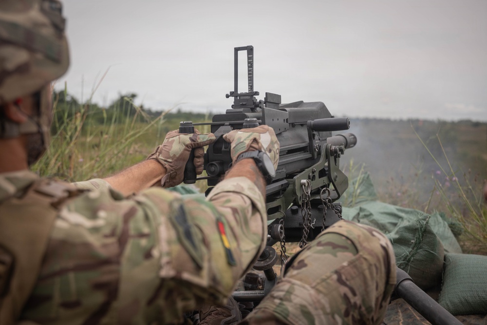 A soldier in multicam camouflage uniform is operating a heavy machine gun from a prone or low position in an open field. The weapon appears to be firing, as evidenced by smoke visible to the right side of the frame. The soldier wears a patch with black, red, and yellow stripes on the left sleeve, consistent with a German flag patch.