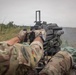 A soldier in multicam camouflage uniform is operating a heavy machine gun from a prone or low position in an open field. The weapon appears to be firing, as evidenced by smoke visible to the right side of the frame. The soldier wears a patch with black, red, and yellow stripes on the left sleeve, consistent with a German flag patch.