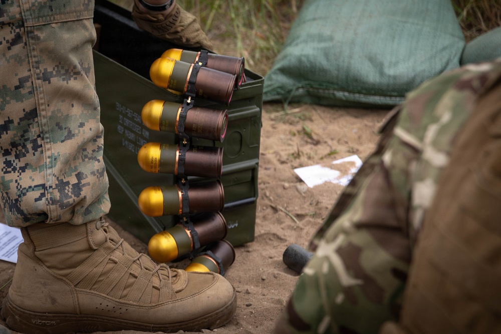 The image shows military personnel in a field setting handling what appear to be 40mm grenade rounds with distinctive gold-colored noses, stored in a standard military ammunition can. At least two individuals are visible, one wearing USMC MARPAT digital camouflage and tan combat boots, the other in a MultiCam pattern uniform. The setting appears to be an outdoor training or operational environment with sandy soil and vegetation in the background.