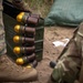 The image shows military personnel in a field setting handling what appear to be 40mm grenade rounds with distinctive gold-colored noses, stored in a standard military ammunition can. At least two individuals are visible, one wearing USMC MARPAT digital camouflage and tan combat boots, the other in a MultiCam pattern uniform. The setting appears to be an outdoor training or operational environment with sandy soil and vegetation in the background.