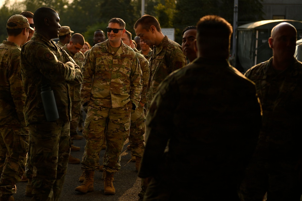 A group of military personnel in OCP (Operational Camouflage Pattern) uniforms are gathered outdoors in what appears to be an informal assembly or meet-and-greet setting. The scene is bathed in warm golden-hour light, suggesting early morning or late evening. A central figure wearing sunglasses appears to be engaged in conversation with fellow service members.