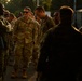 A group of military personnel in OCP (Operational Camouflage Pattern) uniforms are gathered outdoors in what appears to be an informal assembly or meet-and-greet setting. The scene is bathed in warm golden-hour light, suggesting early morning or late evening. A central figure wearing sunglasses appears to be engaged in conversation with fellow service members.