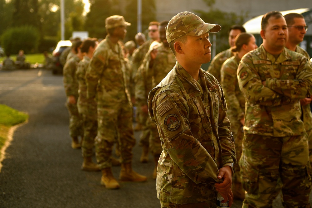 A group of military personnel in OCP (Operational Camouflage Pattern) uniforms are standing in formation or assembly outdoors during golden hour lighting. The central figure, wearing a patrol cap and displaying a US flag patch and unit patch on the left sleeve, appears to be at attention or listening to instructions. The setting appears to be a military installation or base.