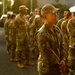 A group of military personnel in OCP (Operational Camouflage Pattern) uniforms are standing in formation or assembly outdoors during golden hour lighting. The central figure, wearing a patrol cap and displaying a US flag patch and unit patch on the left sleeve, appears to be at attention or listening to instructions. The setting appears to be a military installation or base.