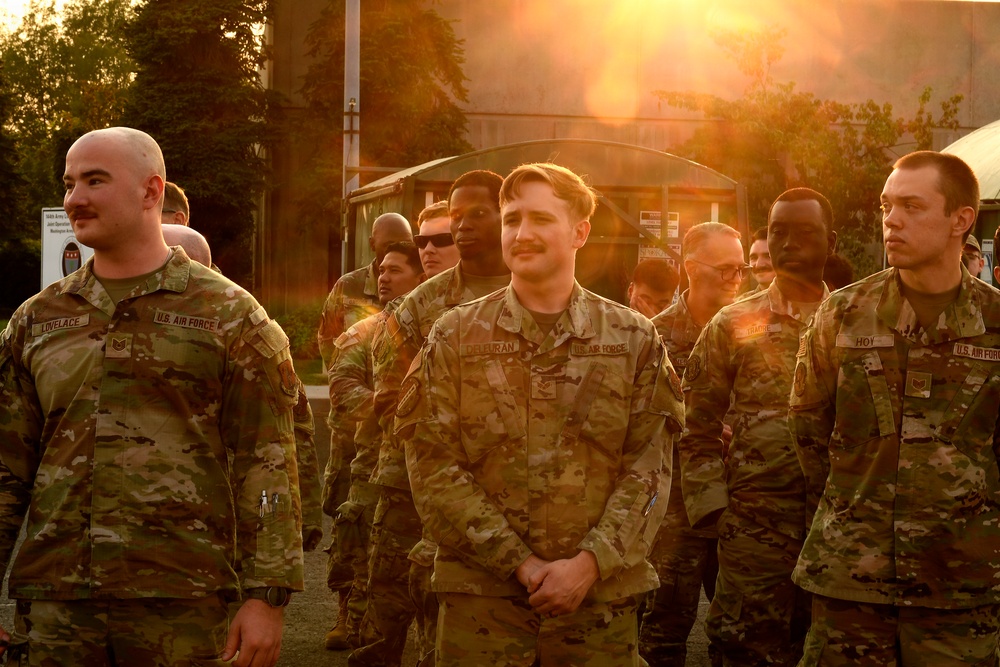 A group of U.S. Air Force personnel in OCP (Operational Camouflage Pattern) uniforms are gathered outdoors during golden hour, with strong sunlight creating a lens flare effect. Visible name tapes include 'LOVELACE' on the left, 'CALHOUN' or similar in the center, and 'HOY' on the right. The personnel appear to be assembled in a formation or group gathering at what appears to be a military installation.