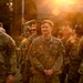 A group of U.S. Air Force personnel in OCP (Operational Camouflage Pattern) uniforms are gathered outdoors during golden hour, with strong sunlight creating a lens flare effect. Visible name tapes include 'LOVELACE' on the left, 'CALHOUN' or similar in the center, and 'HOY' on the right. The personnel appear to be assembled in a formation or group gathering at what appears to be a military installation.