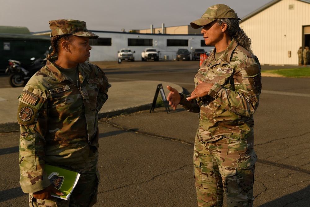 Two female military personnel in OCP (Operational Camouflage Pattern) uniforms are engaged in conversation on what appears to be a military installation or airfield. The individual on the right appears to be speaking and gesturing with her hands, while the one on the left listens and holds a green folder or document. The setting is an outdoor paved area with military vehicles and buildings visible in the background.