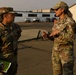 Two female military personnel in OCP (Operational Camouflage Pattern) uniforms are engaged in conversation on what appears to be a military installation or airfield. The individual on the right appears to be speaking and gesturing with her hands, while the one on the left listens and holds a green folder or document. The setting is an outdoor paved area with military vehicles and buildings visible in the background.