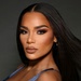 A professional portrait photograph of a woman with long straight black hair, wearing glamorous makeup and a blue top. The image appears to be a high-quality beauty or fashion shoot with studio lighting. The subject is posed facing slightly to the side against a neutral dark gray background.