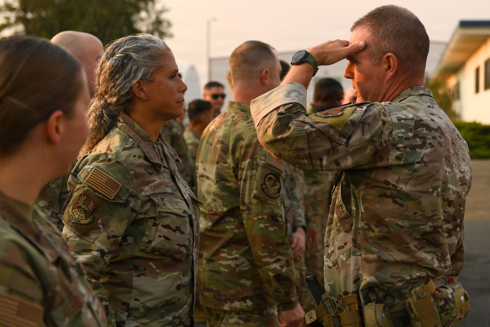 A military formation scene showing uniformed personnel in OCP (Operational Camouflage Pattern) uniforms. A senior officer on the right is rendering a salute toward a female service member, suggesting a formal recognition or inspection event. The setting appears to be an outdoor military installation with hazy atmospheric conditions.