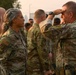 A military formation scene showing uniformed personnel in OCP (Operational Camouflage Pattern) uniforms. A senior officer on the right is rendering a salute toward a female service member, suggesting a formal recognition or inspection event. The setting appears to be an outdoor military installation with hazy atmospheric conditions.