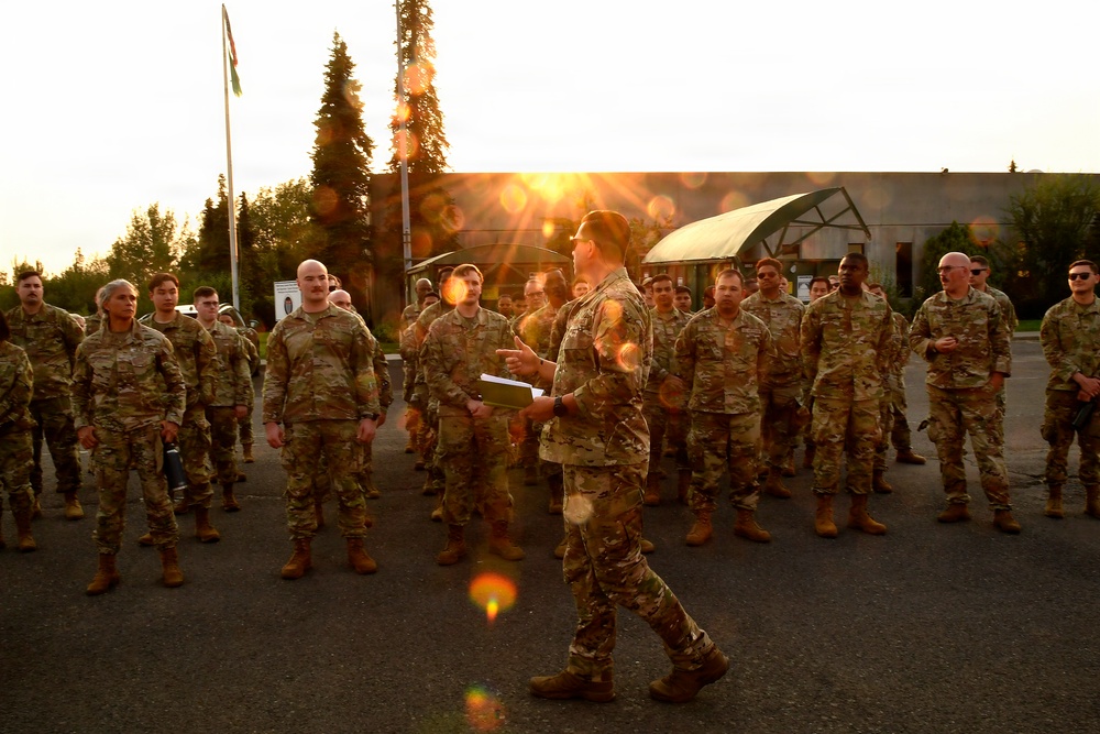 A military officer or NCO is addressing a large formation of soldiers in camouflage uniforms on a paved outdoor area, likely during an early morning or evening briefing. The speaker is holding papers and appears to be conducting a formation brief or roll call. The scene is backlit by a low sun creating lens flare effects, suggesting dawn or dusk conditions.
