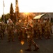 A military officer or NCO is addressing a large formation of soldiers in camouflage uniforms on a paved outdoor area, likely during an early morning or evening briefing. The speaker is holding papers and appears to be conducting a formation brief or roll call. The scene is backlit by a low sun creating lens flare effects, suggesting dawn or dusk conditions.