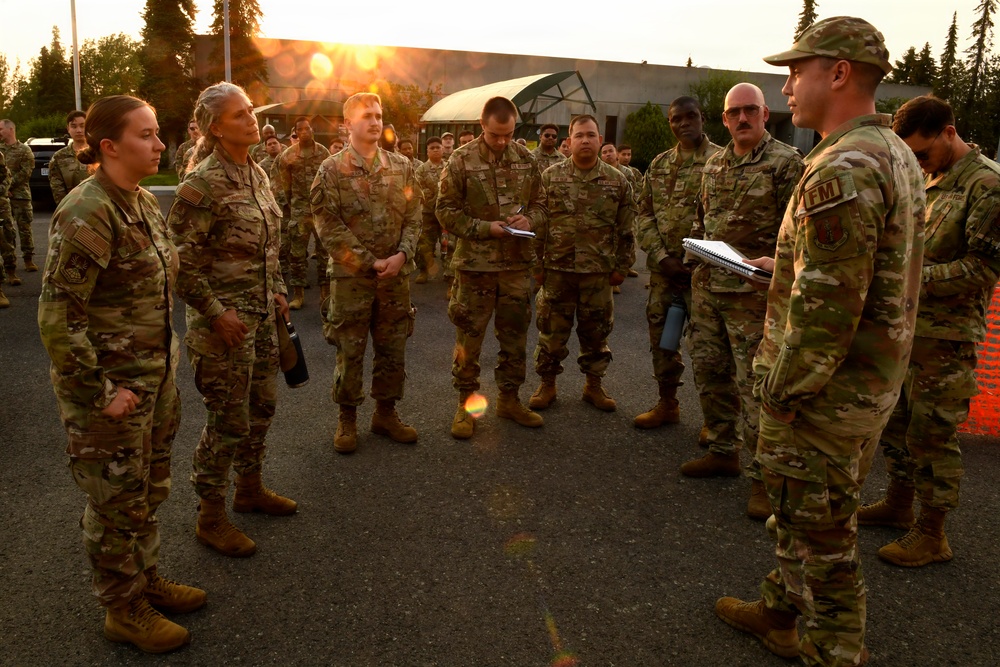 A group of military personnel in OCP (Operational Camouflage Pattern) uniforms are gathered in a semicircle formation during what appears to be an outdoor briefing or formation at dawn or dusk. One soldier on the right, wearing a cap and holding a notebook with a visible 'FM' tab, appears to be addressing the group. The setting appears to be a military installation, likely in a northern or Alaskan environment based on the vegetation and lighting.