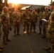 A group of military personnel in OCP (Operational Camouflage Pattern) uniforms are gathered in a semicircle formation during what appears to be an outdoor briefing or formation at dawn or dusk. One soldier on the right, wearing a cap and holding a notebook with a visible 'FM' tab, appears to be addressing the group. The setting appears to be a military installation, likely in a northern or Alaskan environment based on the vegetation and lighting.