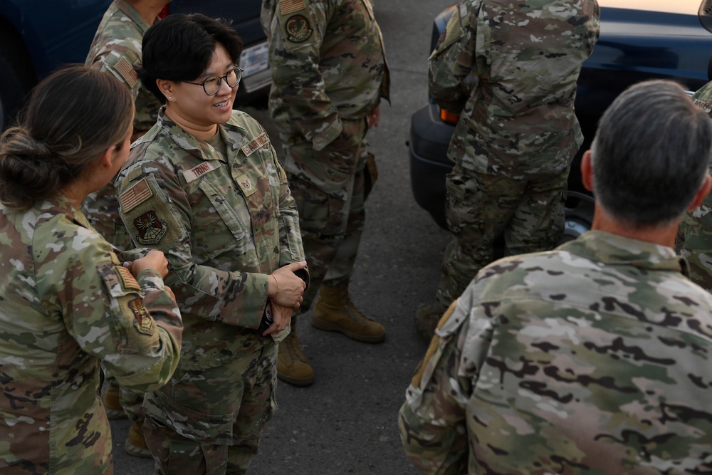 A group of U.S. Air Force personnel in Operational Camouflage Pattern (OCP) uniforms are gathered outdoors, appearing to be in conversation. The central figure, identified by a name tape reading 'TRINH,' is smiling and engaged in discussion with fellow service members. The setting appears to be a parking area or flight line environment.