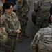 A group of U.S. Air Force personnel in Operational Camouflage Pattern (OCP) uniforms are gathered outdoors, appearing to be in conversation. The central figure, identified by a name tape reading 'TRINH,' is smiling and engaged in discussion with fellow service members. The setting appears to be a parking area or flight line environment.