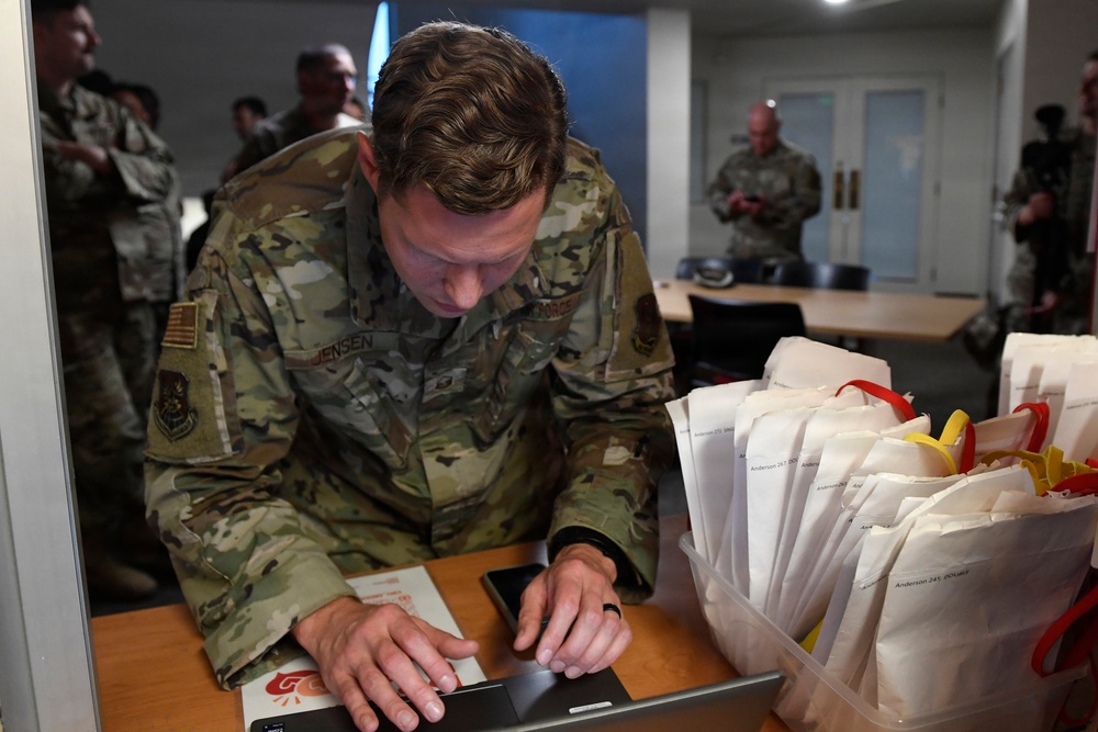 A U.S. Air Force service member named Jensen is leaning over a table, operating a tablet device. Multiple white paper bags labeled with 'Anderson' and numbers are stacked to his right, suggesting a distribution or check-in event. Other uniformed military personnel are visible in the background of what appears to be an indoor facility.