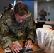 A U.S. Air Force service member named Jensen is leaning over a table, operating a tablet device. Multiple white paper bags labeled with 'Anderson' and numbers are stacked to his right, suggesting a distribution or check-in event. Other uniformed military personnel are visible in the background of what appears to be an indoor facility.