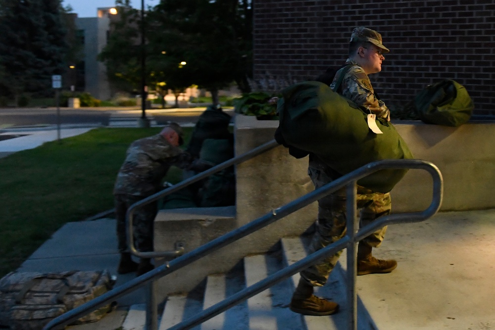 Two military personnel in camouflage uniforms are handling large green duffel bags near the entrance steps of a brick building at dusk or dawn. The foreground soldier is standing on the steps carrying a tagged green bag while looking outward, and a second soldier is bent over near the railing, also handling gear. The setting appears to be a military installation or institutional building with a parking area and trees visible in the background.