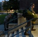 Two military personnel in camouflage uniforms are handling large green duffel bags near the entrance steps of a brick building at dusk or dawn. The foreground soldier is standing on the steps carrying a tagged green bag while looking outward, and a second soldier is bent over near the railing, also handling gear. The setting appears to be a military installation or institutional building with a parking area and trees visible in the background.