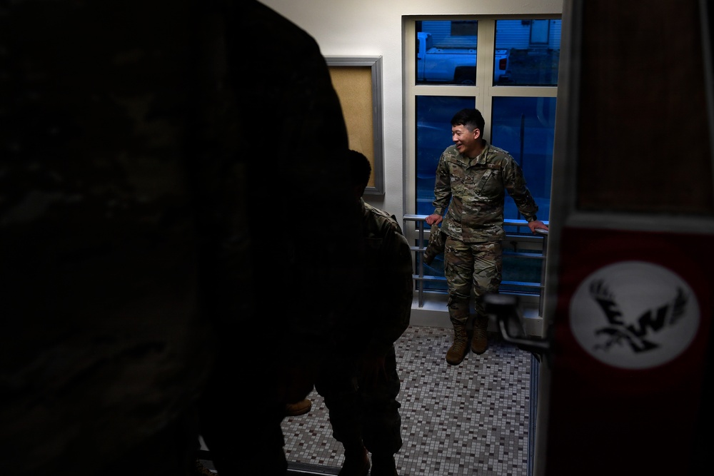The image shows military personnel in camouflage uniforms inside what appears to be a building entrance or lobby area. One soldier in the center is smiling and appears to be engaged in conversation with others. The scene is dimly lit with a bright window providing backlighting.