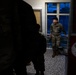 The image shows military personnel in camouflage uniforms inside what appears to be a building entrance or lobby area. One soldier in the center is smiling and appears to be engaged in conversation with others. The scene is dimly lit with a bright window providing backlighting.