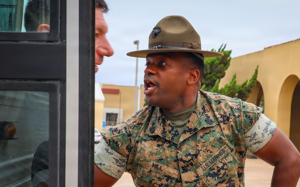 A U.S. Marines drill instructor wearing the distinctive campaign cover (drill sergeant hat) and MARPAT camouflage uniform is shouting at recruits near a bus. His name tape reads 'HILL' and he is in a commanding stance with hands on hips. The setting appears to be a Marine Corps recruit depot, likely MCRD San Diego, based on the architecture and environment.