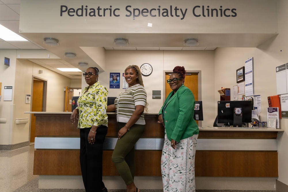 Three women are posing together at the reception desk of a Pediatric Specialty Clinics facility. They appear to be staff members or healthcare professionals, dressed in professional attire and smiling for the photo. The setting is a well-lit clinical reception area with standard medical office furnishings.